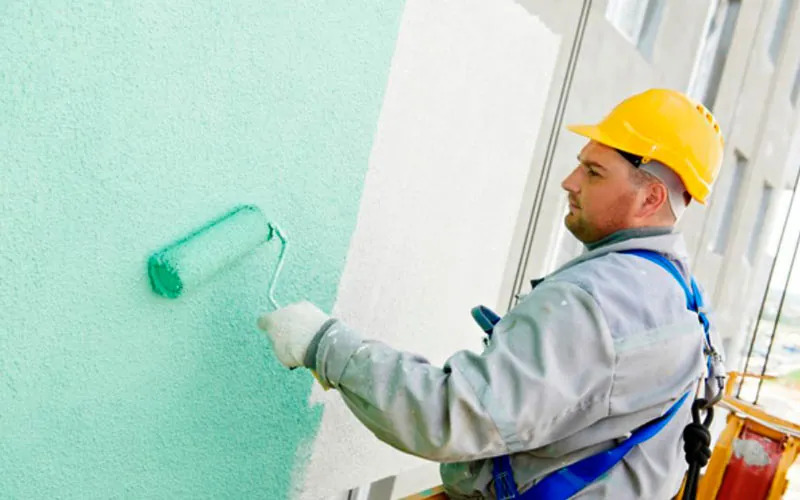 Pintor con casco y arnés aplicando pintura verde agua con rodillo sobre un muro